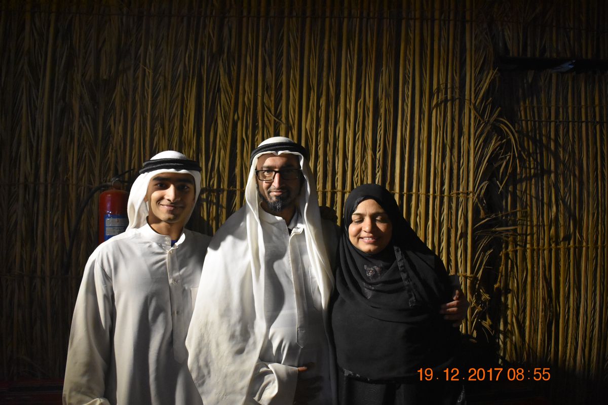 Two men in white kandura and a woman in black abaya by a bamboo wall at night
